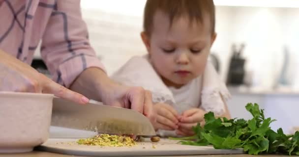 大きなキッチンナイフでカッティングボードにピスタチオを切る女性 小さな娘がママを調理するのを助けます — ストック動画