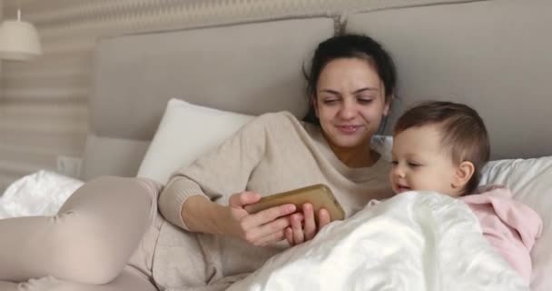 自宅の寝室でベッドに座っている間 スマートフォンで遊ぶ母とかわいい赤ん坊 — ストック動画