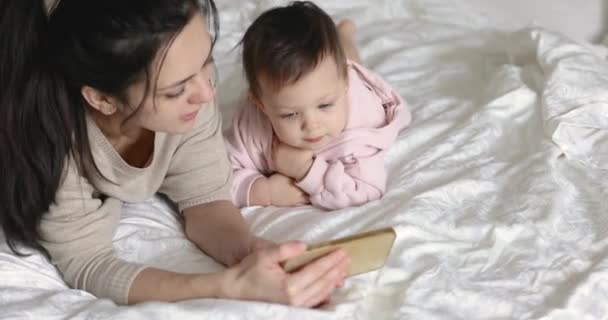 自宅の寝室でベッドに座ってスマートフォンで遊んでいる母と子供の娘 — ストック動画