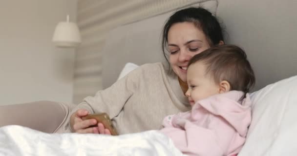 幸せな母親と小さな子供は自宅の寝室でベッドに座っている間スマートフォンで遊んでいます — ストック動画