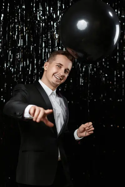 Happy man in black jacket with black air balloons points at camera choosing you on glitter background.