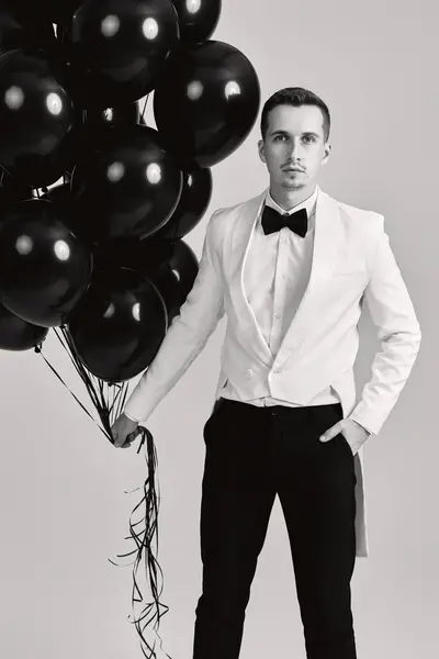 elegant caucasian man in white suit tuxedo with bunch of black air balloons on white background.