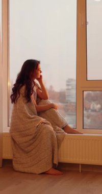 Bir battaniyeye sarılmış gülümseyen kadın pencere eşiğinde oturuyor ve dışarı bakıyor.