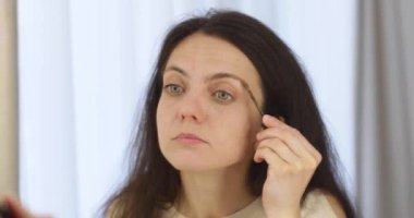 Güzel bir kadın kaşlarını kozmetik fırçayla fırçalıyor ve evde aynaya bakıyor.