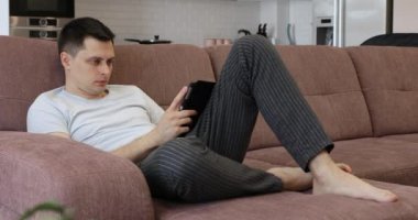 Evdeki kanepede otururken tablet kullanan beyaz bir adam.
