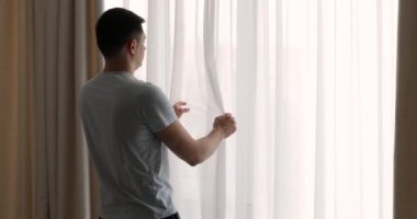 Mavi tişörtlü adam evdeki yatak odasında perdeleri açıyor.