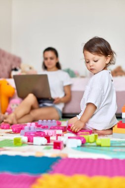 Oyuncak bloklarıyla oynayan küçük tatlı bir kız bebek ve odada dizüstü bilgisayarla yerde oturan bir anne.