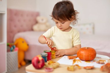 Cadılar Bayramı 'nda balkabağı boyayan mutlu bir kız çocuğu. Gülümseyen bebek sonbahar balkabağını sarı boyayla çiziyor