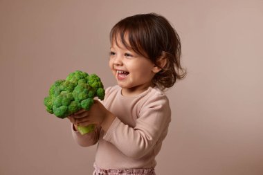 Gülümseyen kız bej arka planda taze yeşil brokoli gösteriyor. sağlıklı bebek maması. Çocuk sebzeleri seviyor