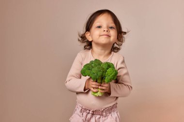 Gülümseyen kız bej arka planda taze yeşil brokoli tutuyor. sağlıklı bebek maması. Çocuk sebzeleri seviyor