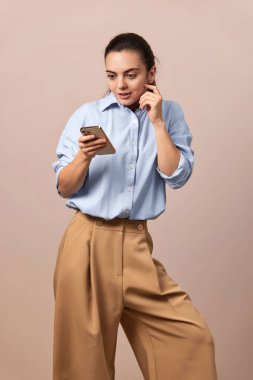 Şok olmuş güzel bir kadın elinde cep telefonuyla bej arka plandaki akıllı telefona bakıyor..