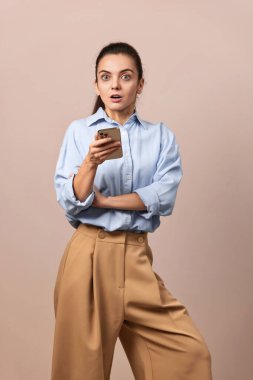 Şok olmuş güzel bir kadın elinde cep telefonuyla bej arka plandaki kameraya bakıyor..