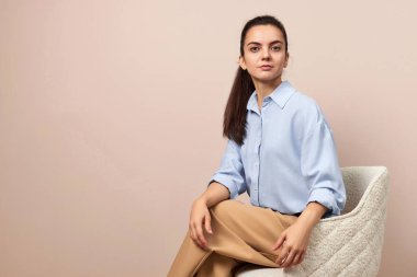 Mavi gömlekli ve bej pantolonlu çekici bir kadın pastel arka planda koltukta oturuyor.