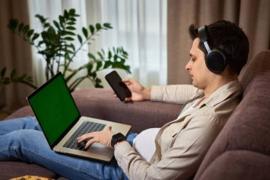 Evdeki koltukta akıllı telefon ve dizüstü bilgisayar kullanan serbest çalışan bir adam.