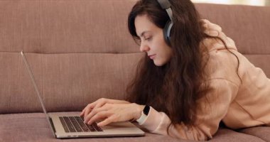 Evdeki kanepede dizüstü bilgisayarlı siyah kulaklıklı bir kadın.