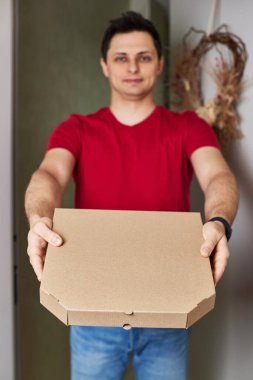 Kırmızı tişörtlü gülümseyen teslimatçı yemek siparişi veriyor, pizza kutusunu kapının önünde tutuyor.