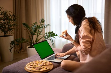 Kafkas kadın dizüstü bilgisayarda çalışıyor ve oturma odasındaki kanepede pizza yiyor.