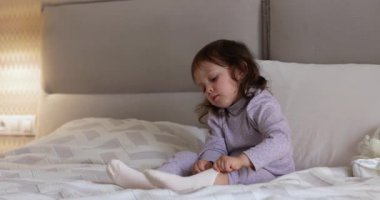 Küçük tatlı kız evde yatakta otururken çıplak ayaklarıyla çorap giymeye çalışıyor.