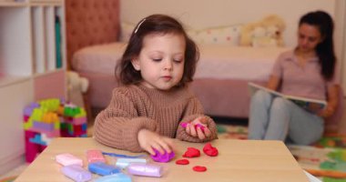 Sevimli küçük kız renkli kille oynuyor ve anne arka planda kitap okuyor. Plastiksin. Hamur oyna