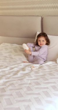 Küçük tatlı kız evde yatakta otururken çıplak ayaklarıyla çorap giymeye çalışıyor.