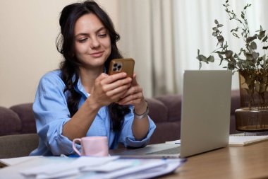 Mavi gömlekli güzel bir kadın ev ofisinde dizüstü bilgisayar kullanıyor.