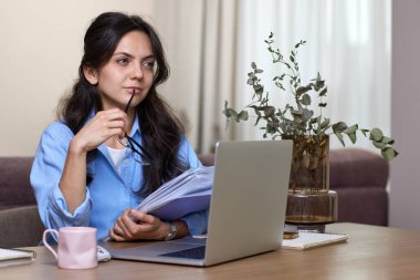 Mavi gömlekli kadın portresi klasörler tutuyor. Ev ofisinde dizüstü bilgisayar kullanıyor.