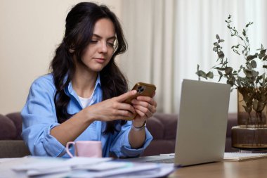 Mavi gömlekli kadın ev ofisinde dizüstü bilgisayar kullanıyor.