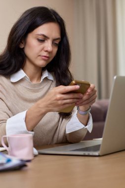 Kadının portresi akıllı telefon tutuyor. Ev ofisinde dizüstü bilgisayar kullanıyor.