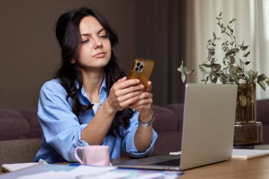 Mavi gömlekli güzel bir kadın ev ofisinde dizüstü bilgisayar kullanıyor.