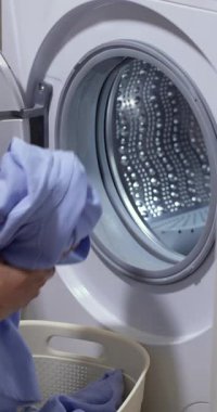 Çamaşır sepetiyle evde çamaşır yıkayan bir kadın. Kadın eli mavi çamaşırları çamaşır makinesinin variline dolduruyor..
