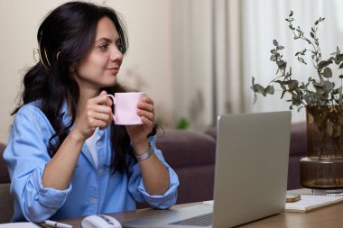 Mavi gömlekli, kahve içen güzel bir kadın ev ofisinde otururken dizüstü bilgisayarda çalışıyor.