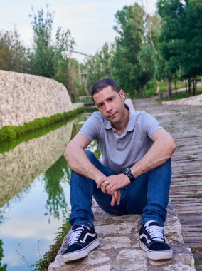 Parkta bir kanalın yanında oturan kot pantolonlu genç bir manken..