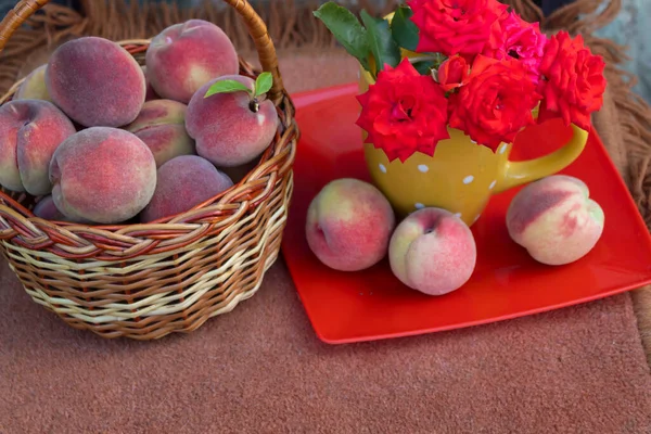 On the table is a basket of ripe peaches and a bouquet of orange roses.