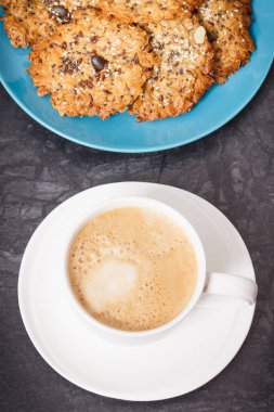 Cup of white coffee and fresh baked homemade oatmeal cookies with honey and different healthy seeds on blue plate. Delicious crunchy dessert