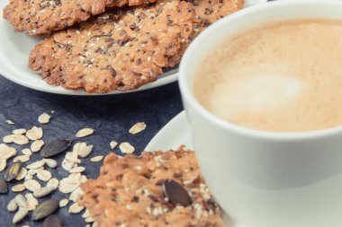 Sütlü bir fincan kahve ve ballı ev yapımı yulaf ezmesi kurabiyeleri ve beyaz tabakta farklı sağlıklı tohumlar. Lezzetli çıtır tatlı.