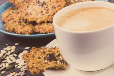 Sütlü bir fincan kahve ve ballı ev yapımı yulaf ezmesi kurabiyeleri ve farklı sağlıklı tohumlar. Lezzetli çıtır tatlı.