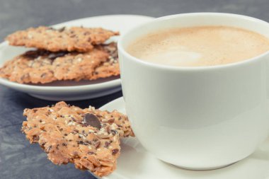 Bir fincan beyaz kahve ve taze ev yapımı yulaf ezmeli kurabiyeler ve farklı sağlıklı tohumlar. Lezzetli çıtır tatlı.