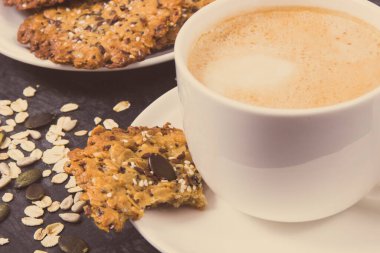 Bir fincan beyaz kahve ve taze ev yapımı yulaf ezmeli kurabiyeler ve farklı sağlıklı tohumlar. Lezzetli çıtır tatlı.