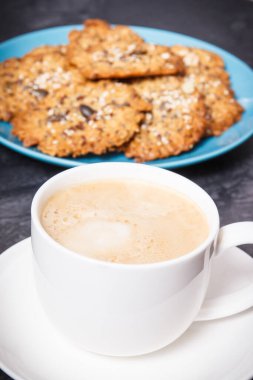 Cup of coffee with milk and fresh baked homemade oatmeal cookies with honey and different healthy seeds on blue plate. Delicious crunchy dessert