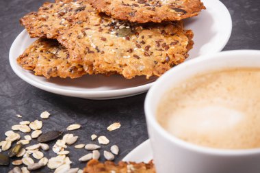 Bir fincan beyaz kahve ve taze ev yapımı yulaf ezmeli kurabiyeler ve farklı sağlıklı tohumlar. Lezzetli çıtır tatlı.