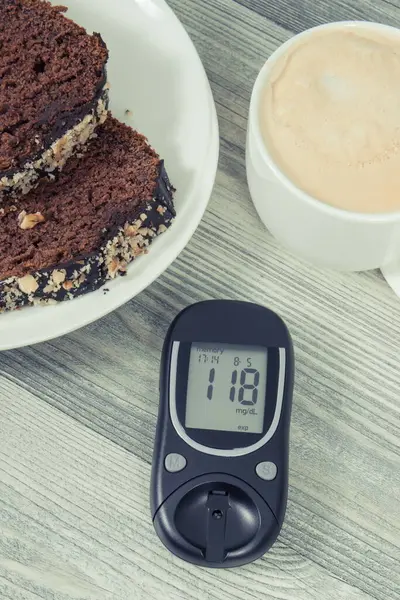Glucometer with result of measurement sugar level, sweet chocolate cake and cup of coffee with milk. Nutrition during diabetes concept