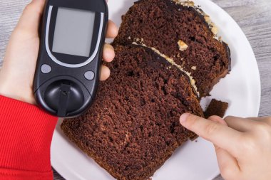 Glikoz ölçer ve çikolatalı pasta tutan bir çocuk eli. Diyabet sırasında şeker seviyesini ölçmek ve kontrol etmek