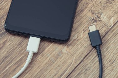 Ahşap arka planda şarj kablosu olan siyah akıllı telefon. Cep telefonu şarj kavramı