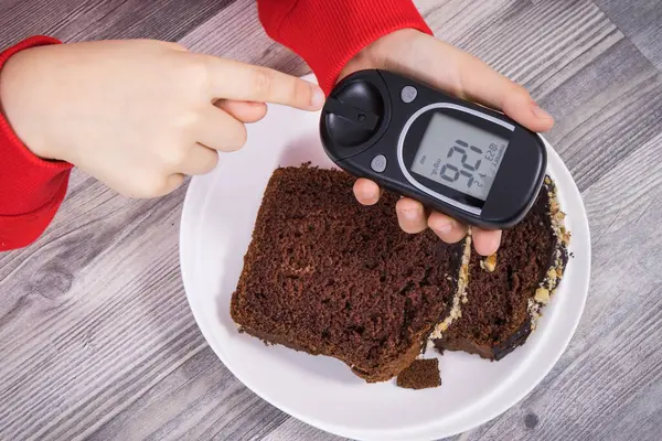 Hand of child holding glucose meter with result of measurement sugar level, sweet chocolate cake. Nutrition during diabetes concept