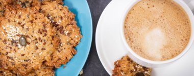 Sütlü bir fincan kahve ve ballı ev yapımı yulaf ezmesi kurabiyeleri ve farklı sağlıklı tohumlar. Lezzetli çıtır tatlı.