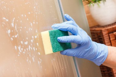 Deterjan ve sünger kullanarak cam duşu temizleyen koruyucu eldivenli yaşlı kadının eli. Ev işleri kavramı