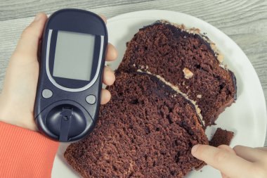 Elinde glukometre, çikolatalı pasta tutan bir çocuk eli. Diyabet sırasında şeker seviyesini ölçmek ve kontrol etmek