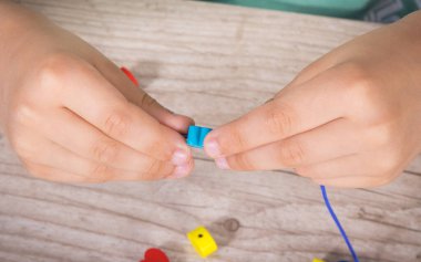 Anaokulu öğrencisi iplikten ve tahta renkli boncuklardan bilezik yapıyor. Çocukların motor becerilerinin, koordinasyonun, yaratıcılığın ve mantıksal düşüncenin geliştirilmesi