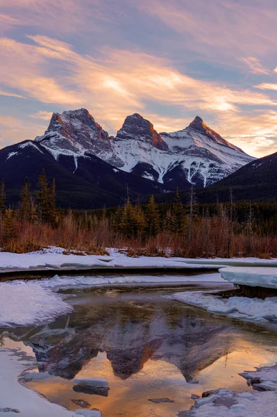 加拿大艾伯塔省坎莫尔附近一座由三座山峰组成的三座山峰在 三姐妹 的金色冬日落日里 高山上的白日余晖在前方的冰面上反射出来 免版税图库图片