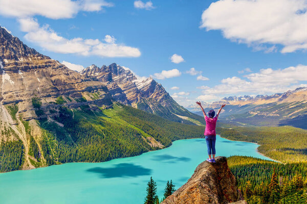 Woman stands on a rock pinnacle of Bow Summit with stunning view of Peyto Lake in the Canadian Rockies of Banff National Park. The glacier-fed lake is famous for its bright turquoise colored waters in the summer.